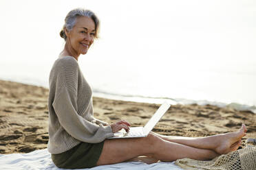 Smiling woman with laptop sitting at beach - EBSF03222