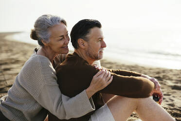 Smiling woman embracing man sitting at beach - EBSF03224