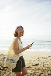 Happy woman with mesh bag holding smart phone at beach - EBSF03236