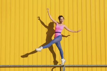 Happy woman doing ballet on railing in front of yellow wall - SYEF00357