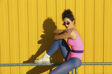 Woman wearing sunglasses sitting on railing in front of yellow wall - SYEF00359