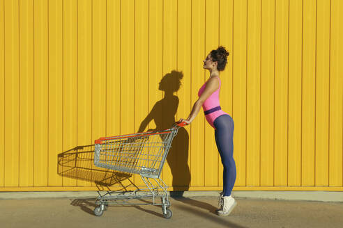 Ballet dancer standing with shopping cart in front of yellow wall - SYEF00361