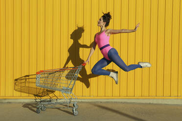 Balletttänzerin springt mit Einkaufswagen vor gelber Wand - SYEF00362
