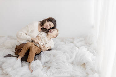Smiling woman tickling son sitting in front of wall - GMLF01442