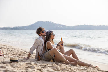 Happy couple enjoying drinks sitting on sand at beach - IKF00188