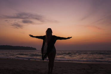 Silhouette einer Frau, die mit ausgestreckten Armen am Strand steht - IKF00196