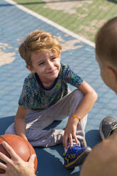 Lächelnder Sohn sitzt mit seinem Vater auf dem Sportplatz - IKF00206