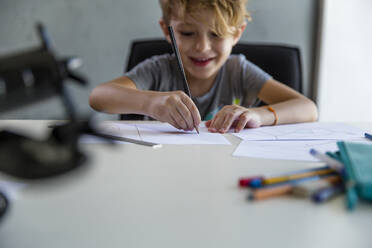 Lächelnder Junge, der mit einem Bleistift am Schreibtisch auf Papier zeichnet - IKF00216