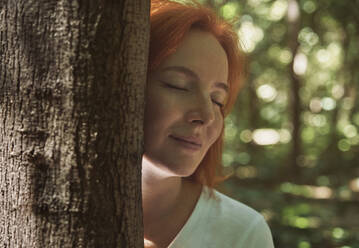 Redhead woman with eyes closed embracing tree trunk in forest - AZF00511