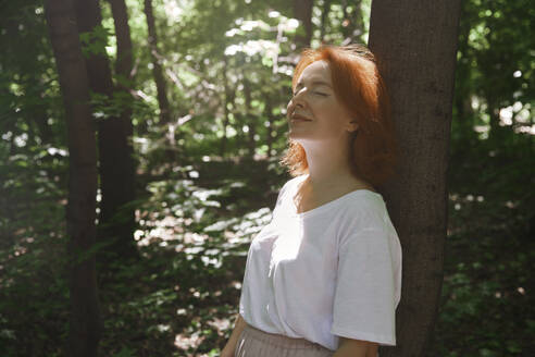 Lächelnde rothaarige Frau mit geschlossenen Augen lehnt an einem Baum im Wald - AZF00513