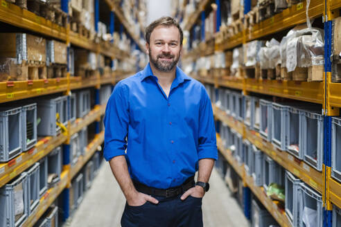 Smiling businessman with hands in pockets standing in warehouse - DIGF19953