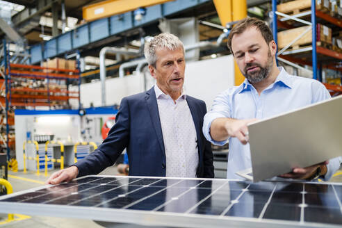 Manager mit Laptop, der von einem Geschäftsmann gehalten wird, der ein Solarpanel in einer Fabrik hält - DIGF20030