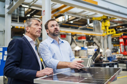 Manager und Geschäftsmann mit Blick auf ein Solarpanel in einer Fabrik - DIGF20032