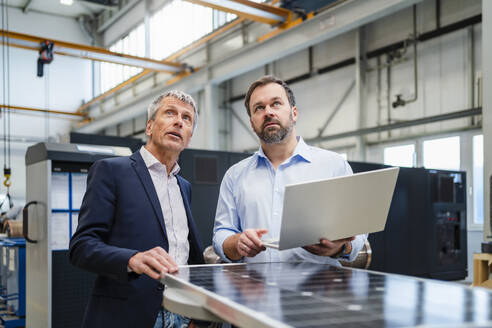 Ein Manager hält einen Laptop in der Hand, während ein Geschäftsmann in einer Fabrik diskutiert - DIGF20034