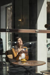 Nachdenkliche Frau mit Smartphone im Café sitzend durch Glas gesehen - JOSEF18606