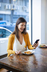Glückliche Frau, die ein Smartphone benutzt, sitzt mit Essen auf dem Tisch in einem Cafe - JOSEF18611
