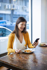 Happy woman using smart phone sitting with food on table at cafe - JOSEF18611