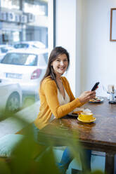 Smiling woman holding smart phone sitting at cafe - JOSEF18612