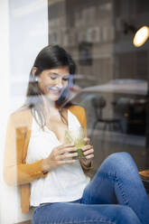 Lächelnde Frau mit gesundem Getränk im Café sitzend durch ein Glasfenster gesehen - JOSEF18629