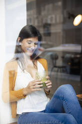 Smiling woman holding healthy drink sitting at cafe seen through glass window - JOSEF18629