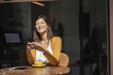 Smiling woman with eyes closed holding smart phone at cafe - JOSEF18640