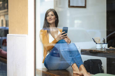 Lächelnde Frau mit Smartphone, die in einem Café sitzt, gesehen durch ein Glasfenster - JOSEF18642
