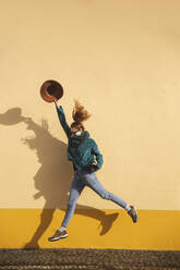 Springende Frau mit Hut und Kamera vor gelber Wand - PCLF00416