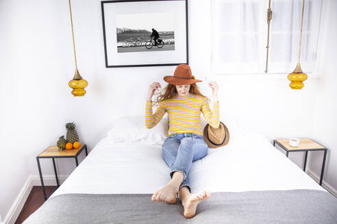 Frau mit Hut sitzt auf dem Bett in einem Hotelzimmer - PCLF00422