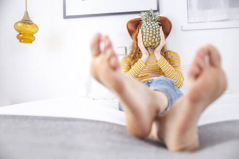 Frau hält Ananas vor dem Gesicht auf dem Bett - PCLF00423