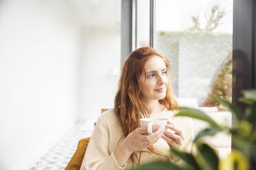 Thoughtful woman sitting with cup in hand by window - PCLF00433