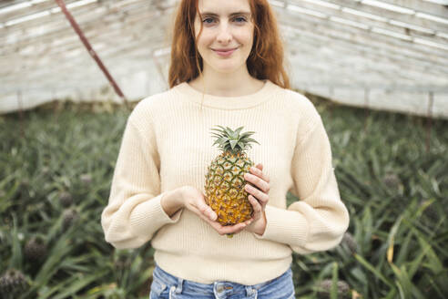 Smiling redhead woman holding pineapple in greenhouse - PCLF00452