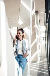 Smiling businesswoman talking on smart phone leaning on column at office - MOEF04541