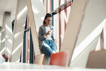 Geschäftsfrau, die ein Smartphone benutzt und sich im Büro an eine Säule lehnt - MOEF04571