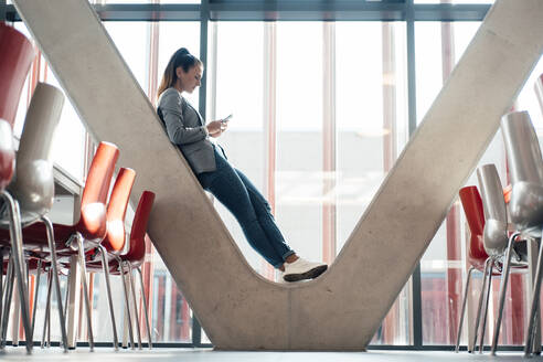 Businesswoman surfing net through smart phone leaning on office column - MOEF04572