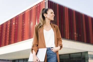 Smiling businesswoman with hand in pocket standing in front of modern building - MOEF04583