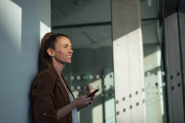 Thoughtful businesswoman holding smart phone leaning on wall at office - MOEF04595