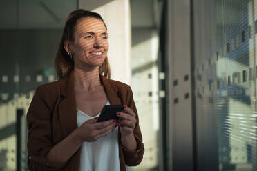 Happy businesswoman holding smart phone at office - MOEF04598