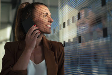 Geschäftsfrau, die mit einem Smartphone spricht und aus dem Fenster im Büro schaut - MOEF04600