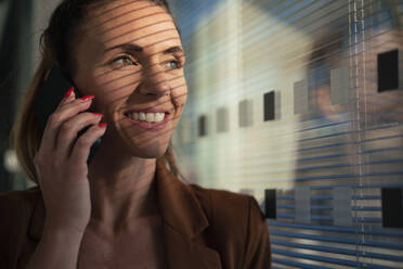Glückliche Geschäftsfrau, die in der Nähe der Wand im Büro mit einem Smartphone spricht - MOEF04601