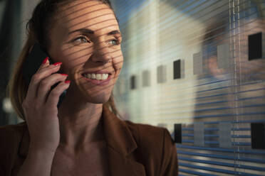 Happy businesswoman talking on smart phone near wall at office - MOEF04601