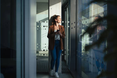 Businesswoman holding smart phone leaning on column at office - MOEF04605