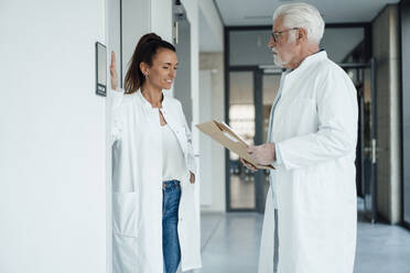 Smiling doctor discussing with senior colleague holding document in hospital - MOEF04616