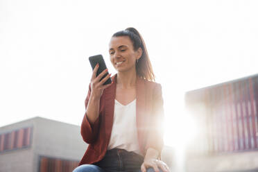 Lächelnde Geschäftsfrau Textnachrichten über Smartphone an einem sonnigen Tag - MOEF04619