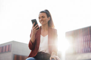 Lächelnde Geschäftsfrau Textnachrichten über Smartphone an einem sonnigen Tag - MOEF04619