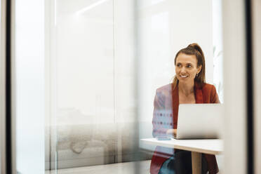 Contemplative businesswoman with laptop sitting at desk seen through glass wall - MOEF04623