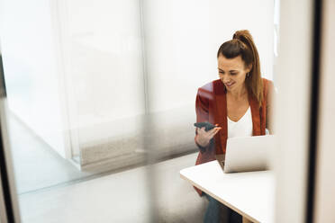 Businesswoman with laptop using smart phone sitting at desk in office - MOEF04625