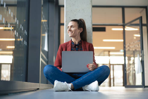 Lächelnde Geschäftsfrau mit Laptop im Schneidersitz auf dem Boden im Büro - MOEF04627