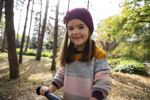 Lächelndes Mädchen mit Strickmütze hält Fahrradgriff im Park - IKF00237