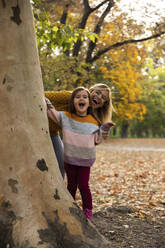 Fröhliche Mutter und Tochter stehen hinter einem Baum - IKF00251