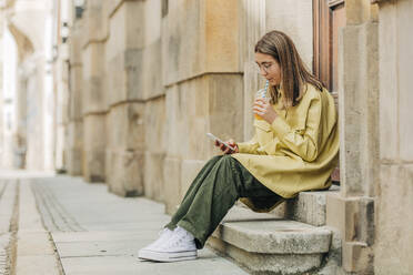 Teenage girl drinking juice and using smart phone sitting on steps - VSNF00718